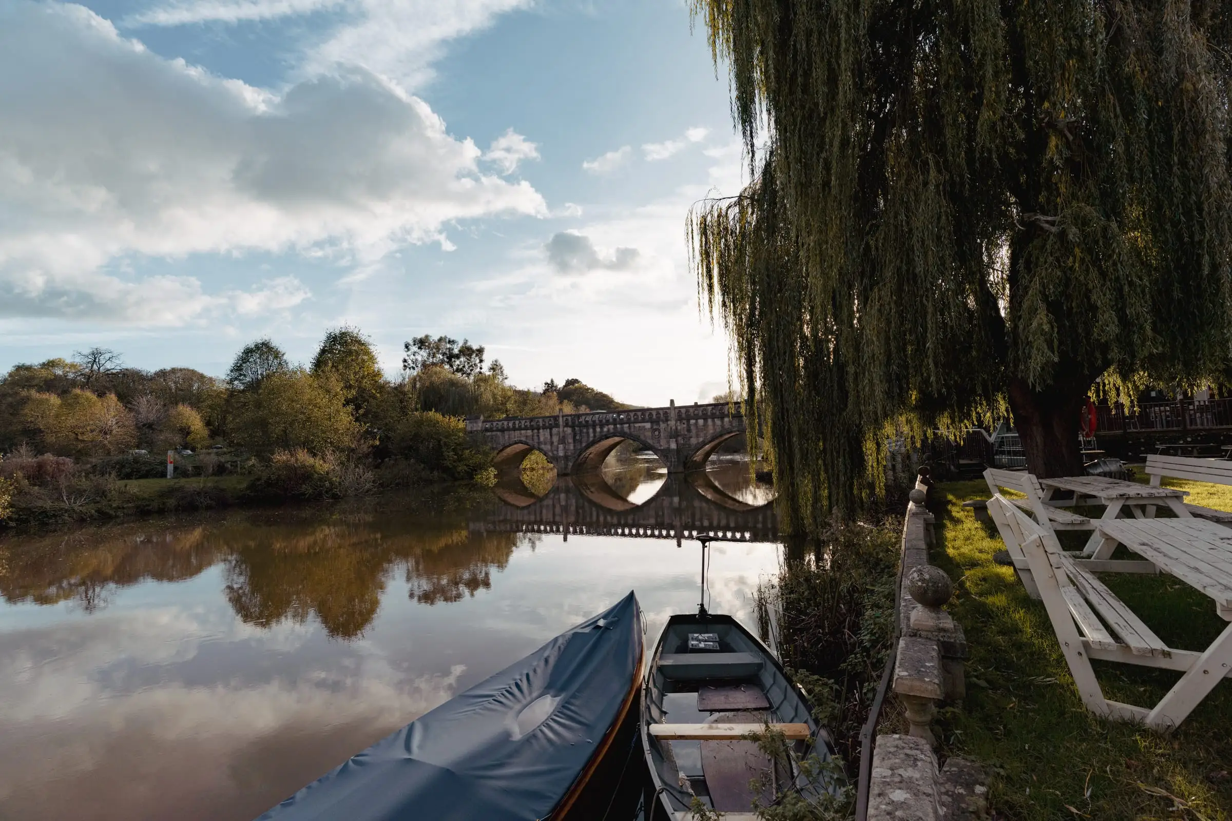 Picnic tables on the lawn by the Avon, under a willow, with the stone bridge reflected in the water